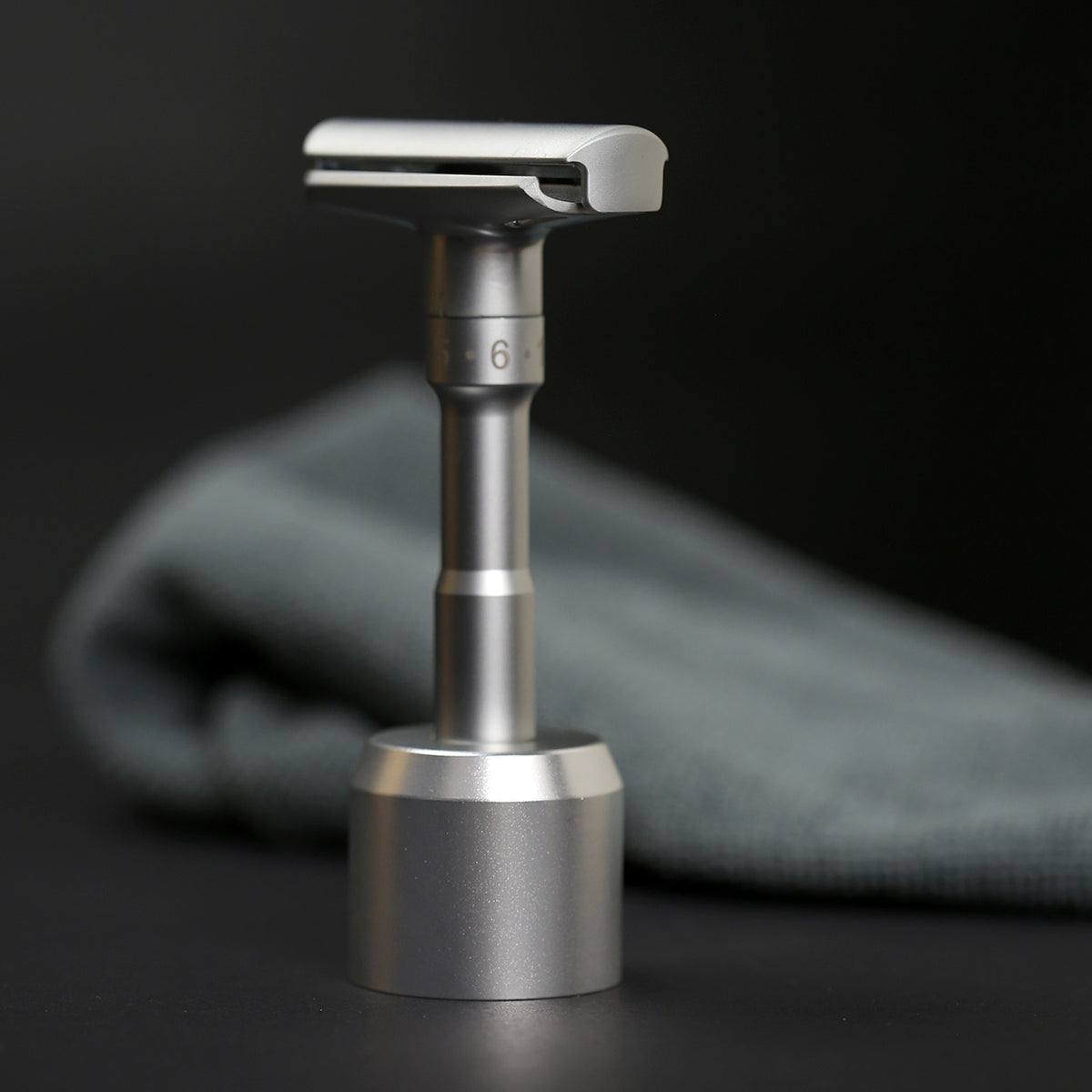 Adjustable saftey razor showing the number 6 setting, in its matching matt chrome stand in a dark setting with dark grey towel.