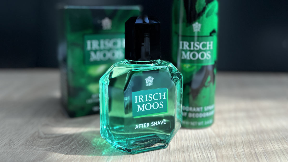 A bottle of Irisch Moos Aftershave which is bright emerald green, on a wooden surface with the box and some Irisch Moos deodorant in the back of the shot