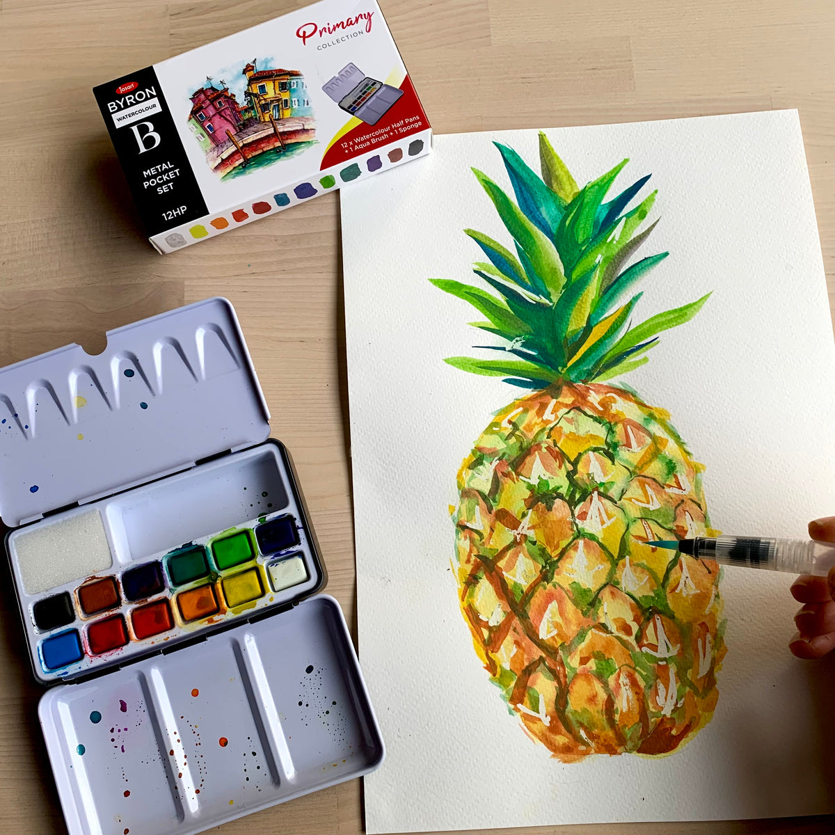 Watercolor painting of a pineapple on paper with a watercolor set and paint box in the background.