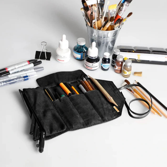 Collection of art supplies including brushes, ink bottles, and a black roll-up case on a light gray background.