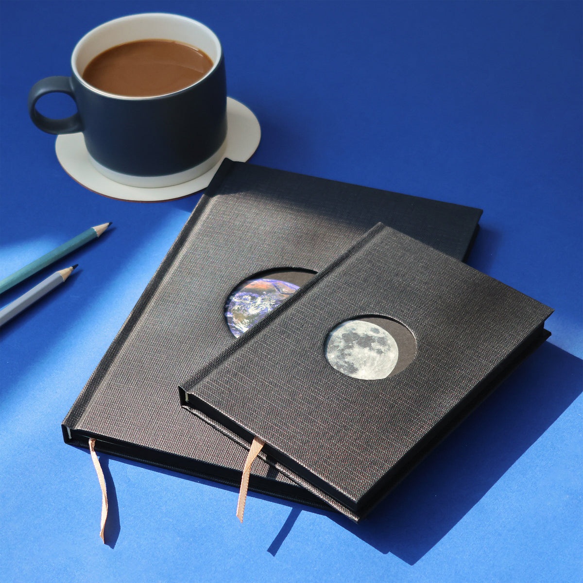 Two black fabric-bound books with celestial designs on a blue surface with a cup of coffee.