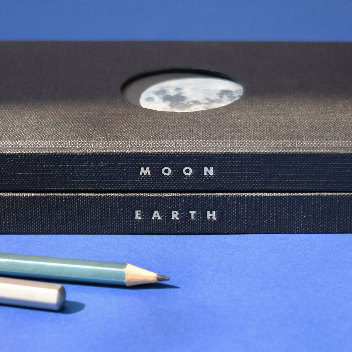 Two notebooks labeled 'Moon' and 'Earth' with a pencil on a blue background