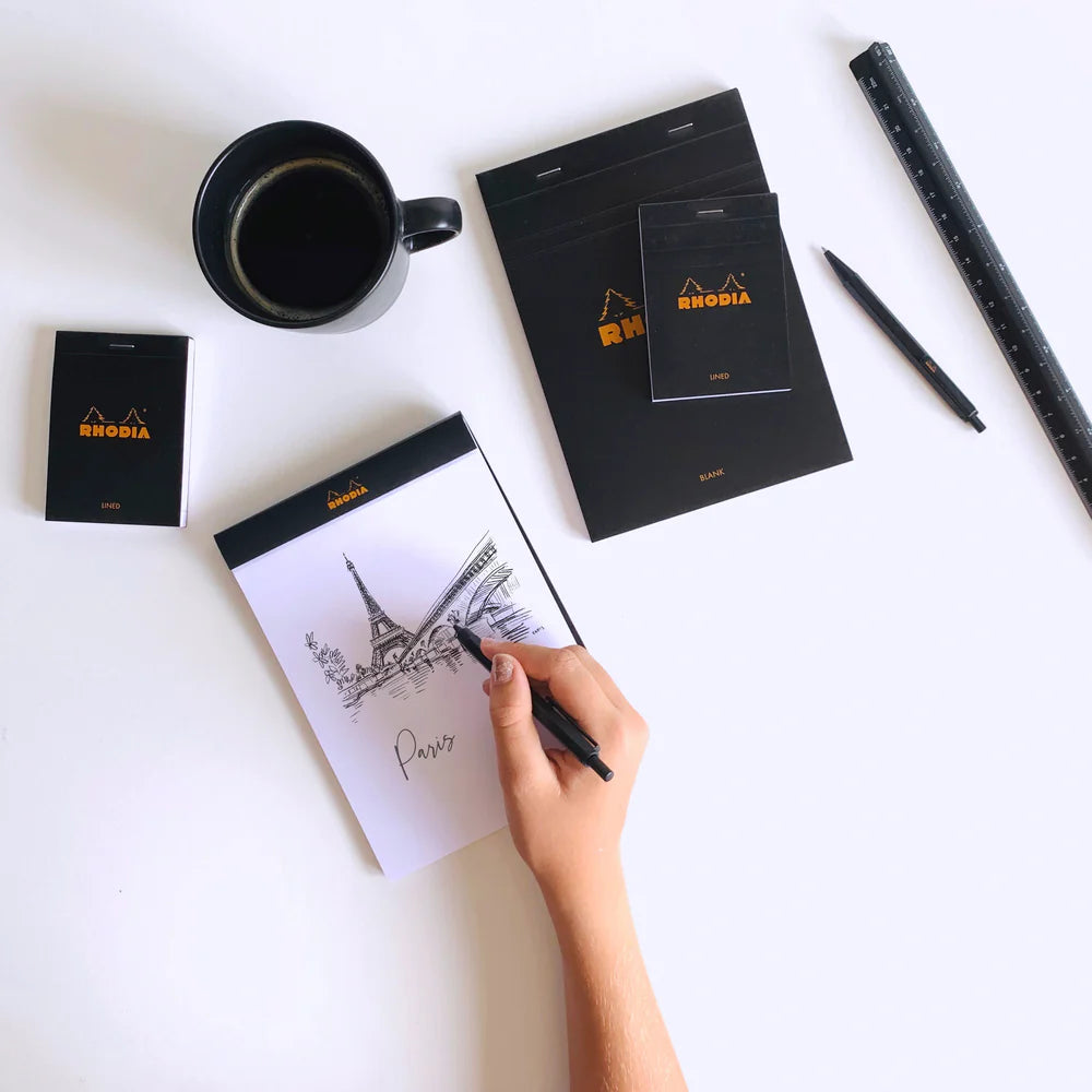 Person drawing the Eiffel Tower on a Rhodia notebook with a cup of coffee and stationery items on a white surface.