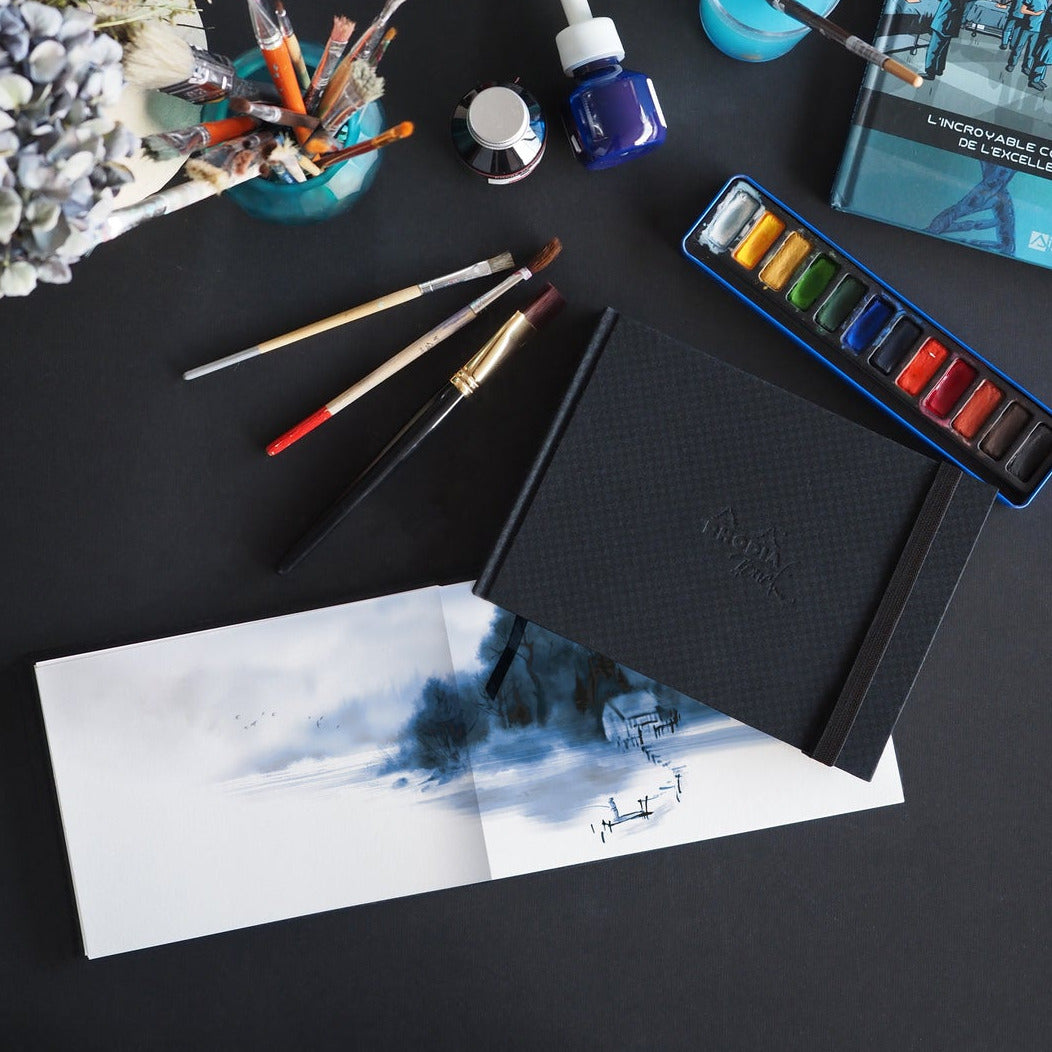 An overhead flat-lay of an art painting set on a dark surface, including a black textured notebook with a blue and white landscape sketch, a blue watercolor paint palette, paintbrushes, a small bottle of blue paint, and other art supplies.