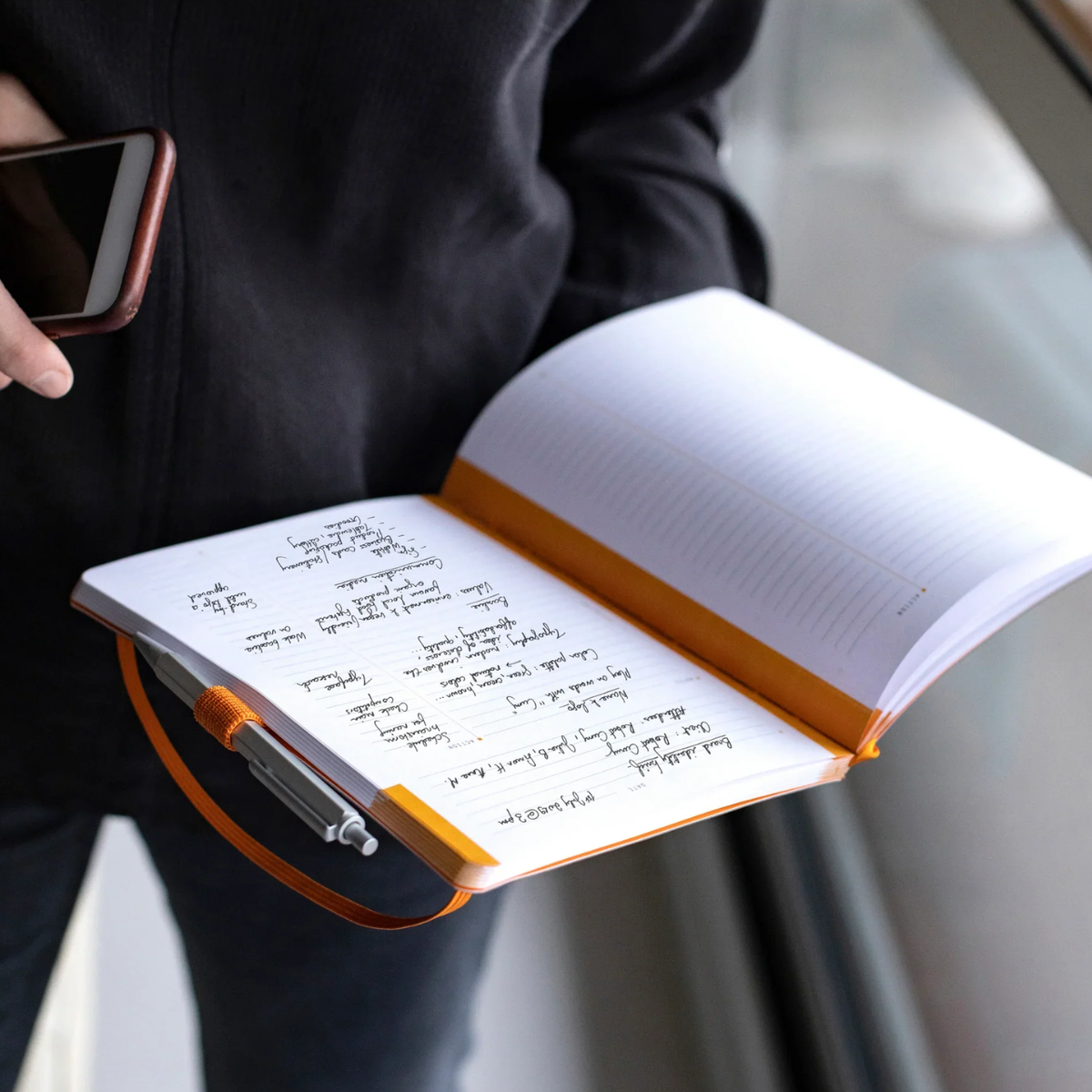 Person holding an open notebook with writing and a smartphone, blurred background