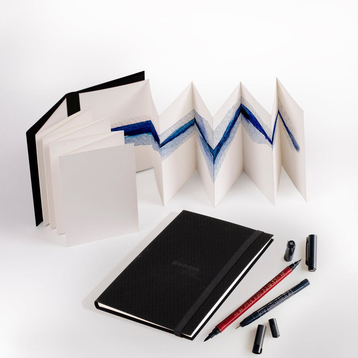 Notebook, pens, and a folded accordion-style book on a white background
