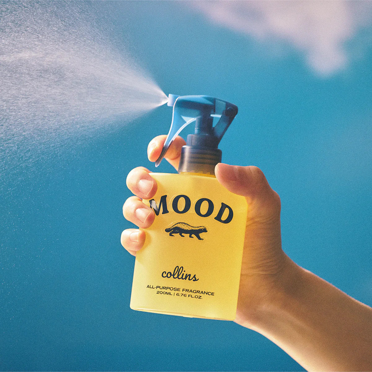 Hand holding a yellow 'Mood' spray bottle against a blue sky background
