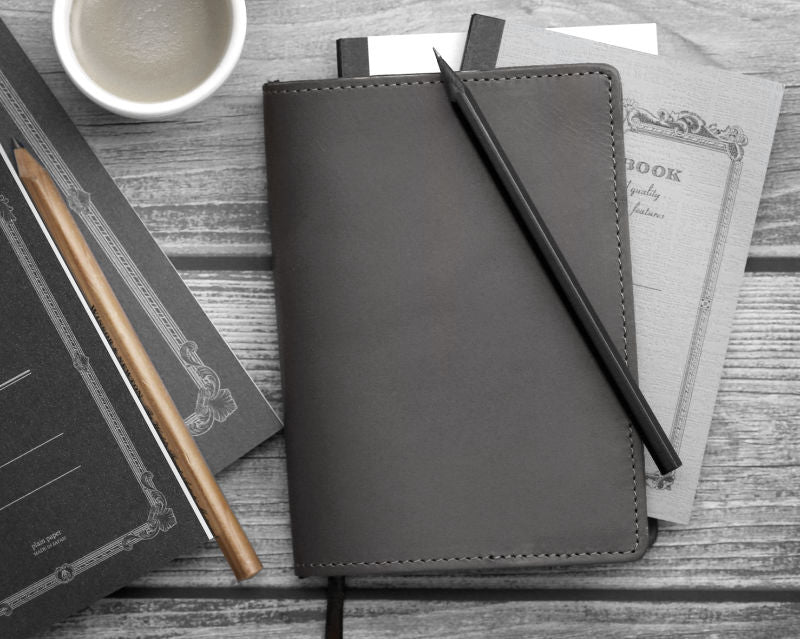 Leather notebook with a pen on a wooden surface with a other notebooks and coffee cup.