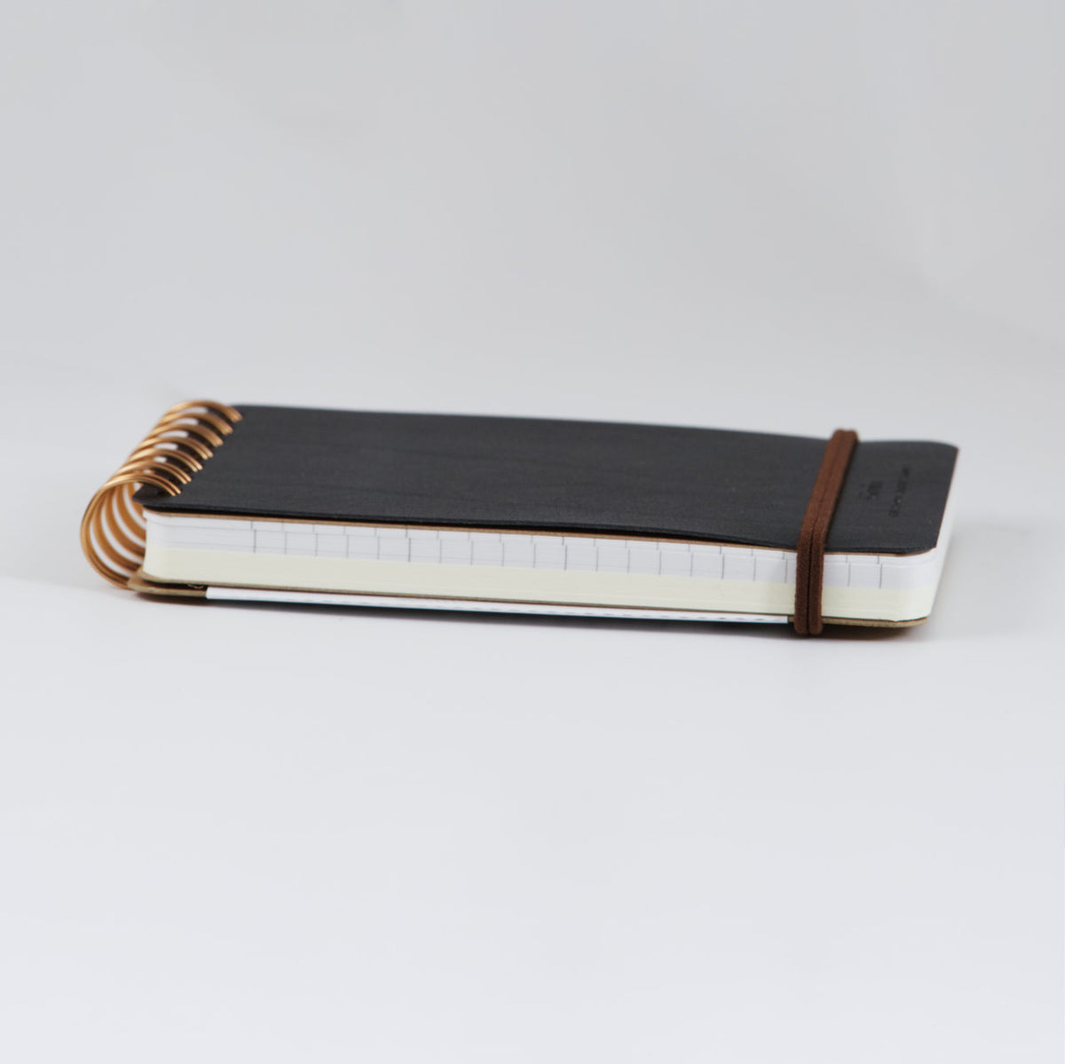 side view of the black memo sized Midori Grain Notebook showing the recycled leather cover, brown double strand elastic closure, Lined Midori paper in the front and blank cream Midori paper in the back 