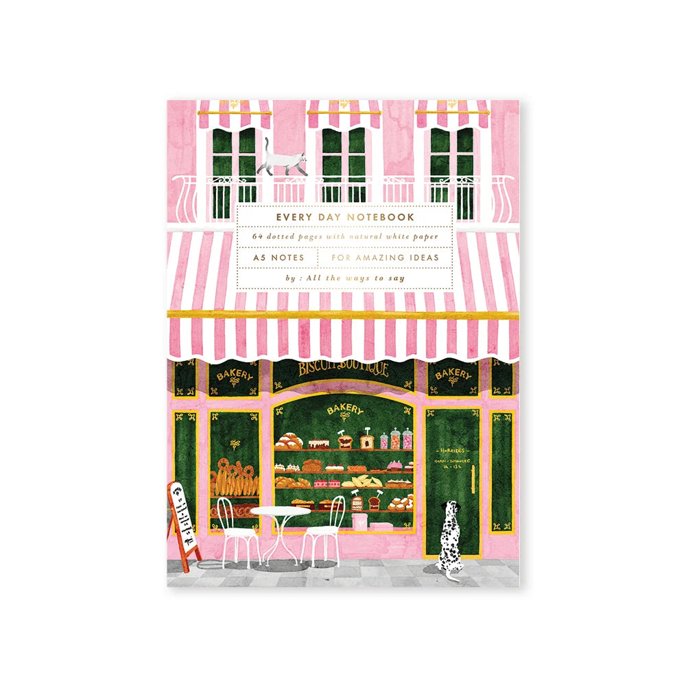 A pink and white striped bakery-themed notebook cover featuring an illustration of a bakery storefront with cafe chairs and a cat, labeled 'Every Day Notebook'.