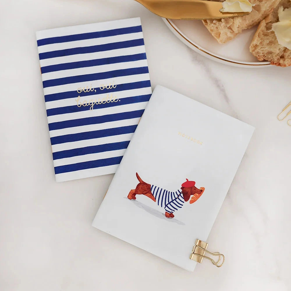 Two decorative notebooks on a white marble surface with a gold butter knife and toast in the background. One notebook has navy and white horizontal stripes with gold foil text reading 'petite baguette'.