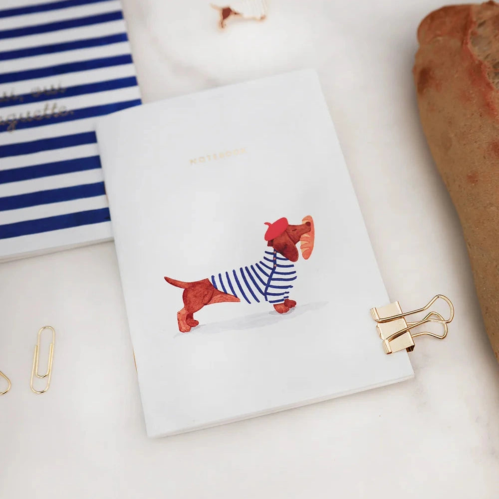 A white notebook with a cartoon dachshund wearing a blue and white striped shirt, styled on a white marble surface with gold paperclips and other stationery props.