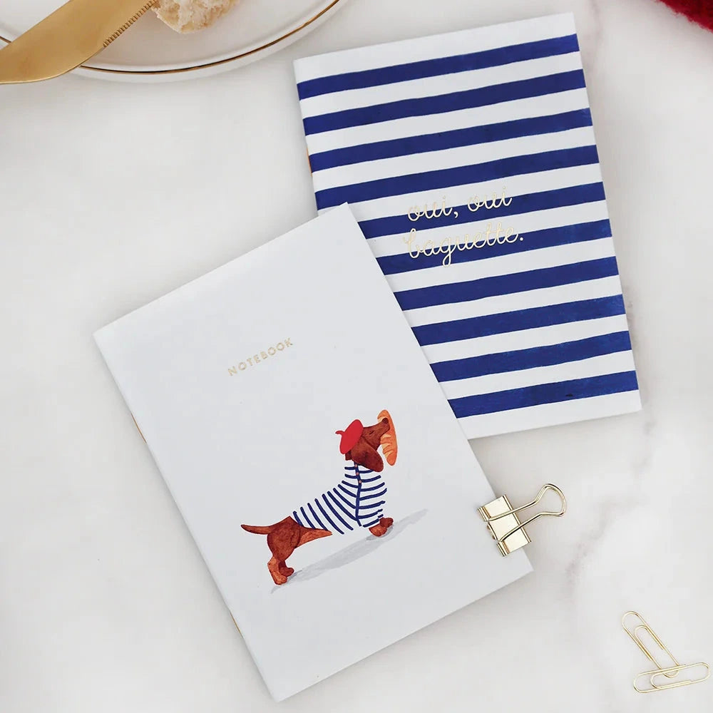 Two small notebooks or cards arranged on a white marble surface with food props. One card is white with a dachshund illustration wearing a navy-and-white striped sweater and the word 'MORTIS' at the top.