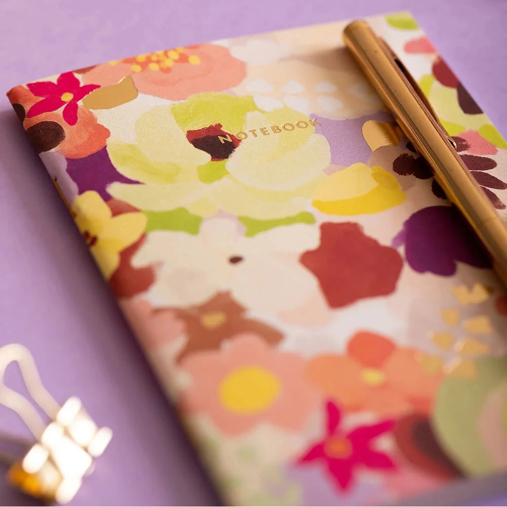 A floral notebook with a gold pen resting on top, placed on a lavender surface with sunglasses and stationery props in the background.