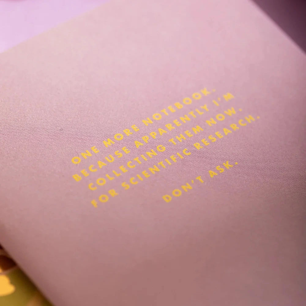 A close-up of a light pink textured notebook with gold foil text on the cover, resting on a lavender surface.