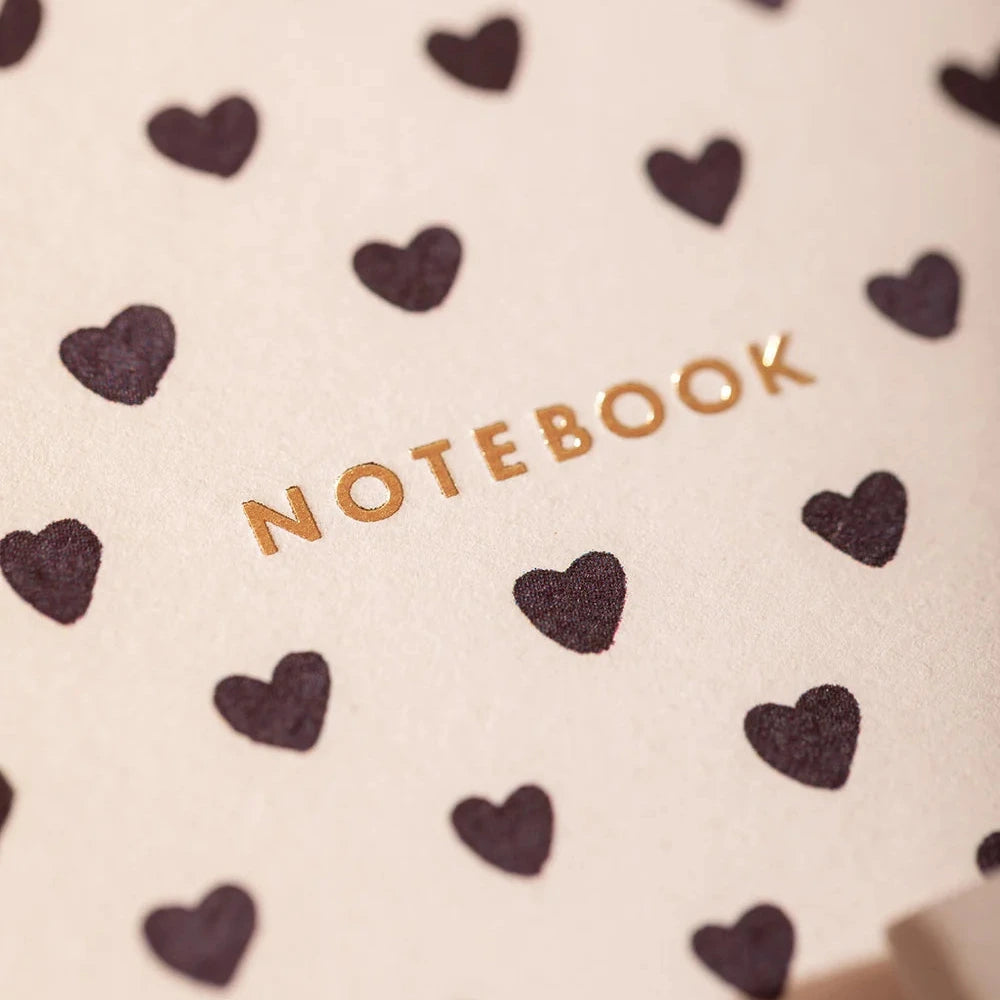 A close-up of a cream-colored notebook cover featuring a repeating pattern of dark brown hearts and the word 'NOTEBOOK' printed in metallic gold foil.