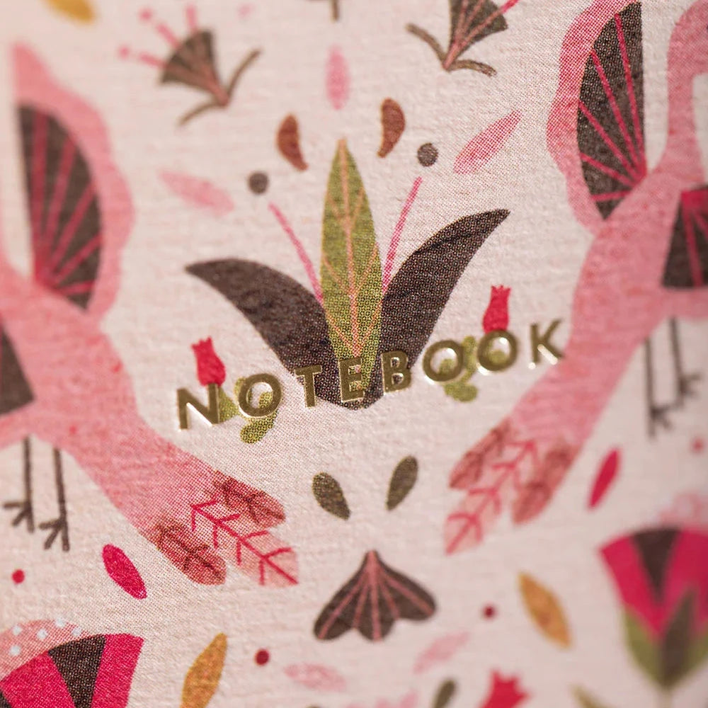 A close-up of a notebook cover featuring a symmetrical floral and botanical pattern in pink, red, green, and brown on a light background, with the word 'NOTEBOOK' embossed in gold across the center.