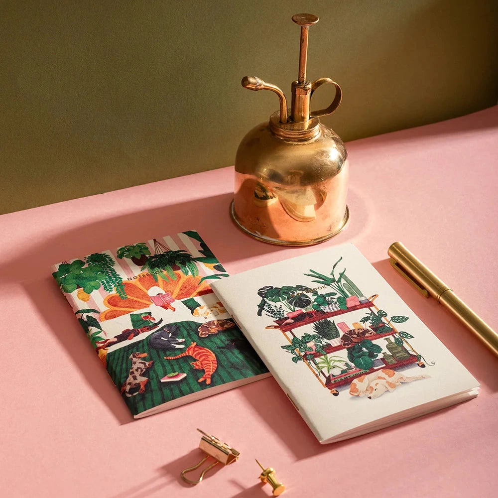 Two notebooks with floral designs on a pink surface with a gold spray bottle.