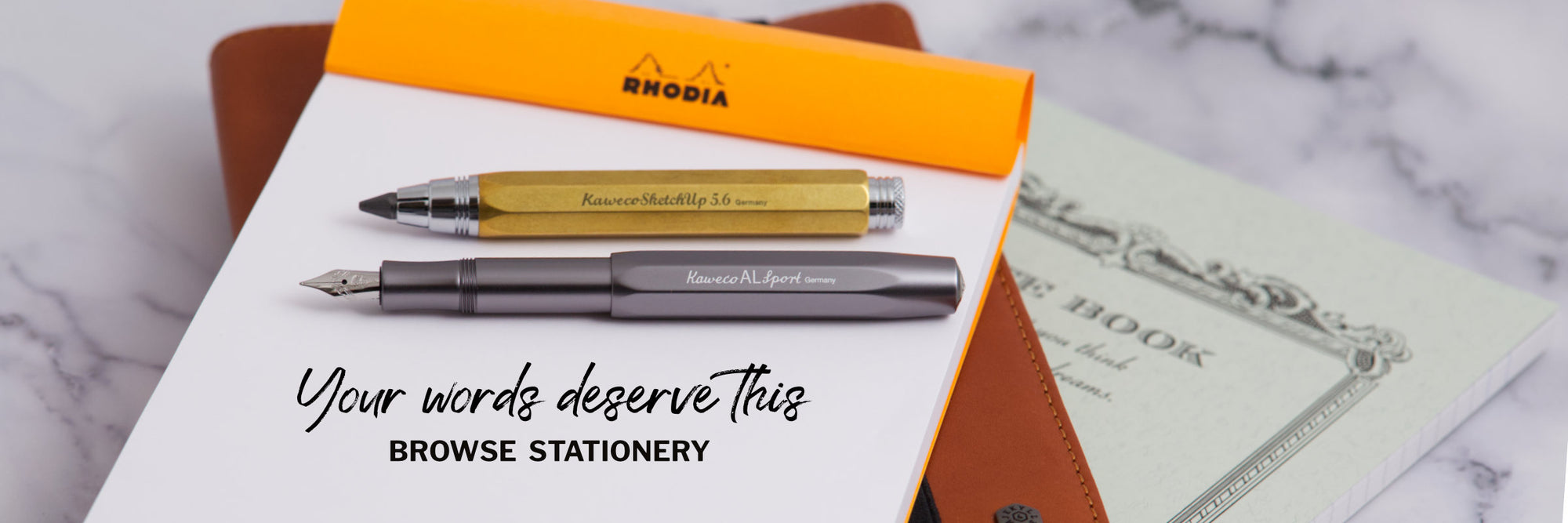 A fountain pen, a clutch pencil and a Rhodia notebook on a marble surface with a leather notebook cover