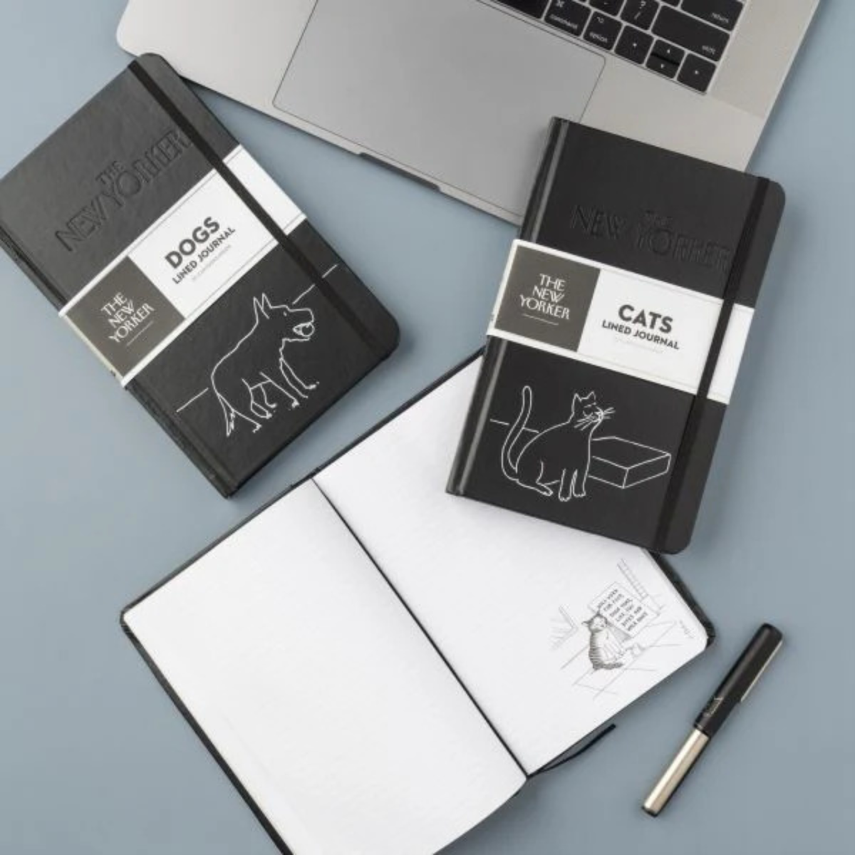 Two black notebooks labeled 'DOGS' and 'CATS' by The New Yorker on a light blue surface with an open notebook and pen.