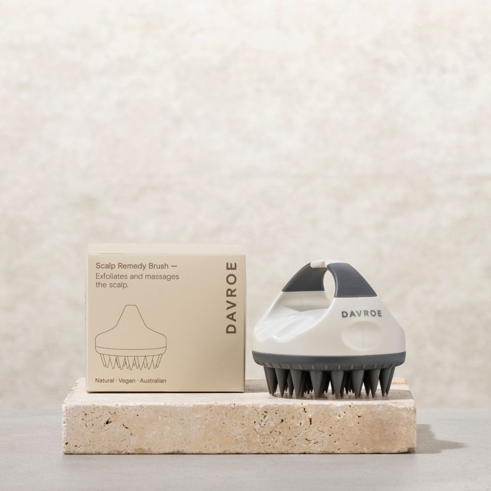 A Davroe Scalp Remedy Brush shown next to its beige packaging box, resting on a textured stone block against a neutral beige background.