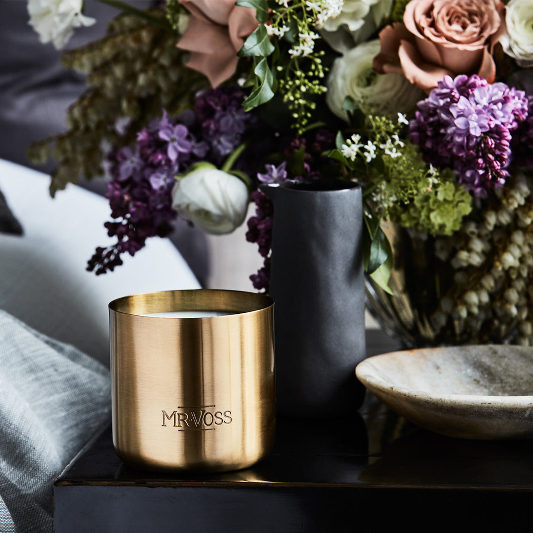 Gold candle with 'MRVOSS' branding in front of a floral arrangement.