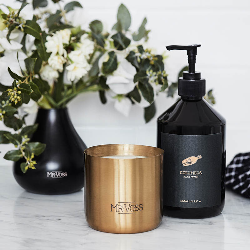 Gold candle and black bottle with 'Mr. Voss' branding on a white surface with flowers in the background.