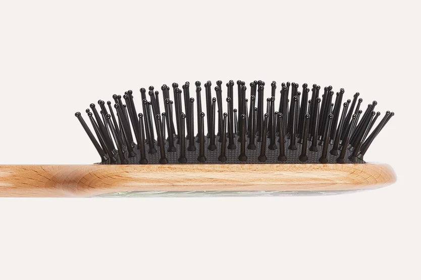A close-up side view of a hairbrush showing a light natural wood handle and black bristles on a plain off-white background.