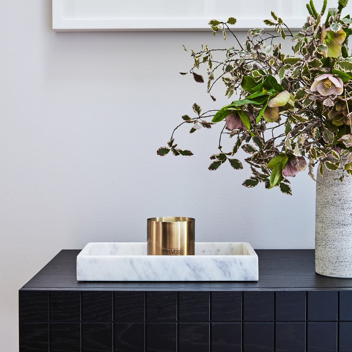 Black tiled table with marble tray, gold candle, and vase with flowers against a white wall.