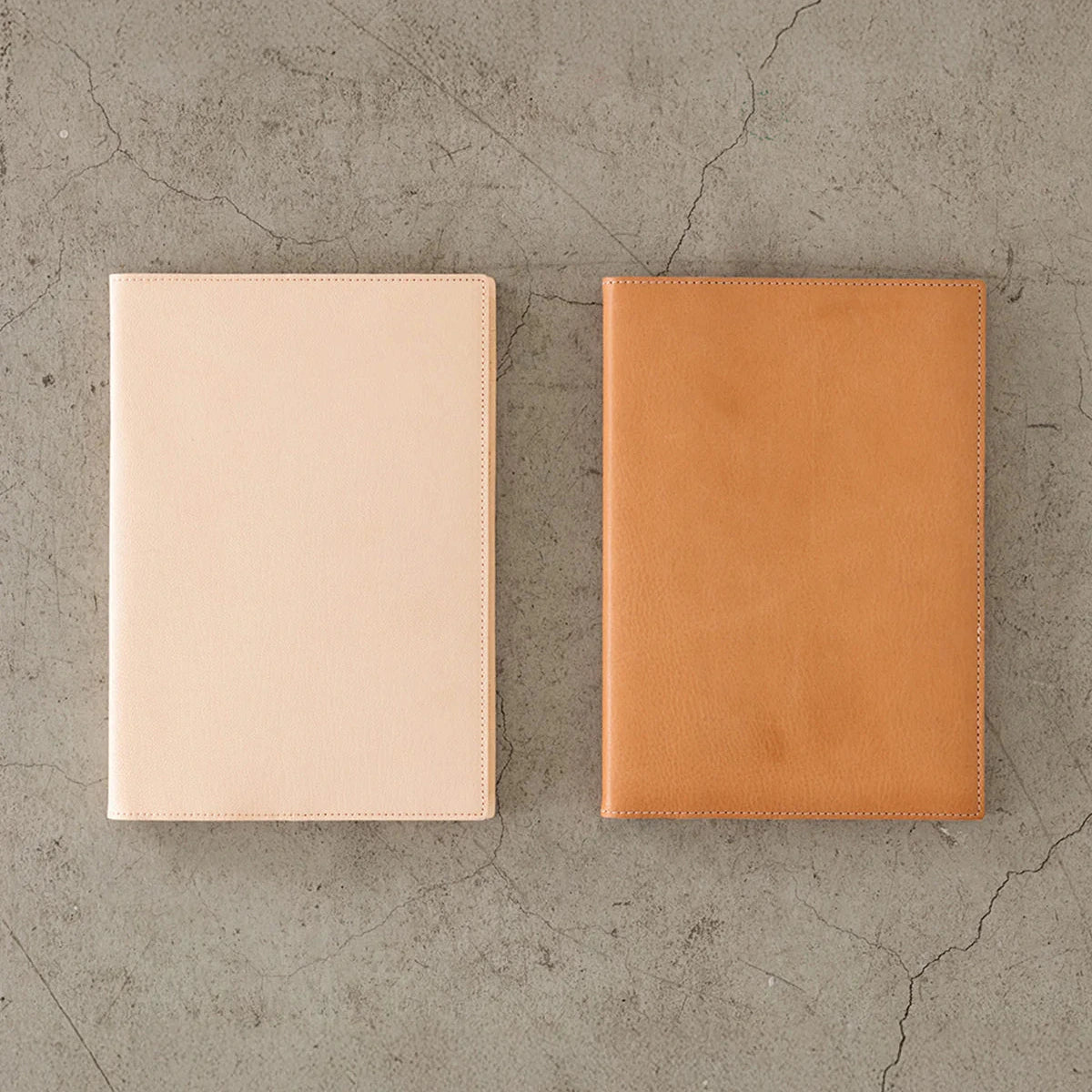 Two leather-bound books, one beige and one brown, on a textured concrete surface.