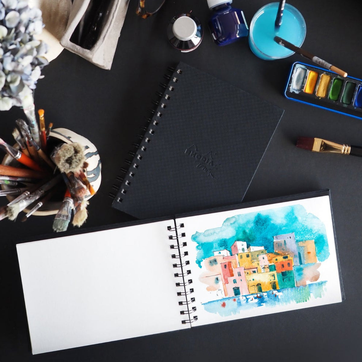 Art supplies including a watercolor painting of a cityscape, ink bottle, and brushes on a black surface.