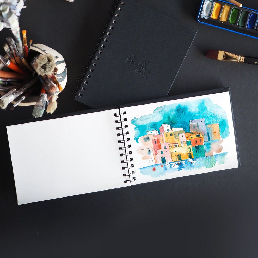 Art supplies including a watercolor painting of a cityscape, ink bottle, and brushes on a black surface.