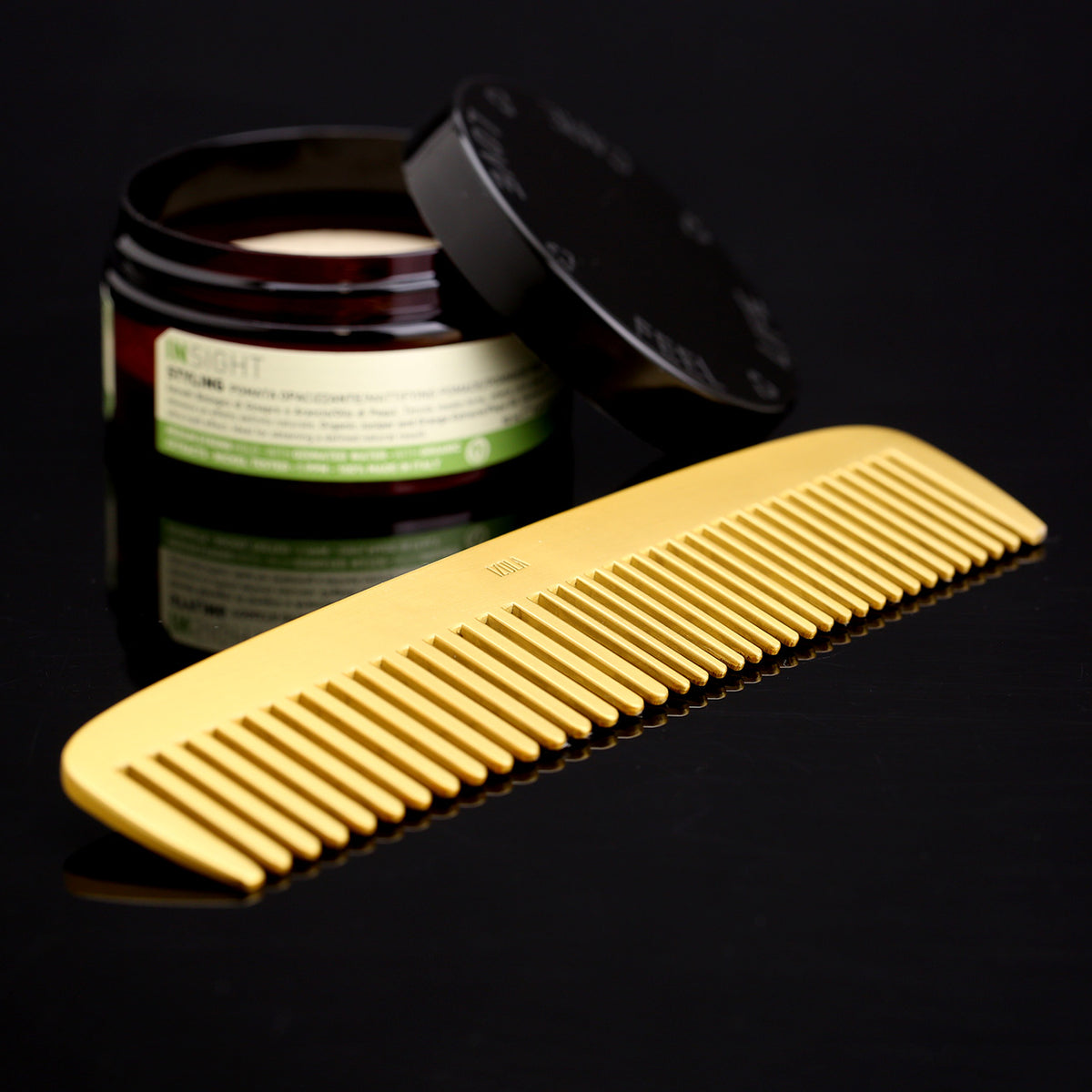 Brass comb on reflective black surface with an open tub of hair pomade for size reference. Pomade sold separately